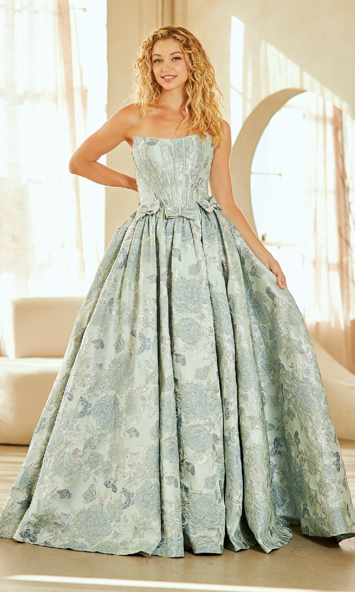 Indoors, a woman smiles while wearing the Lace-Up Print Prom Ball Gown: Amelia Couture 8068—a strapless, full-skirted light blue floral brocade dress—holding part of the gown to the side.